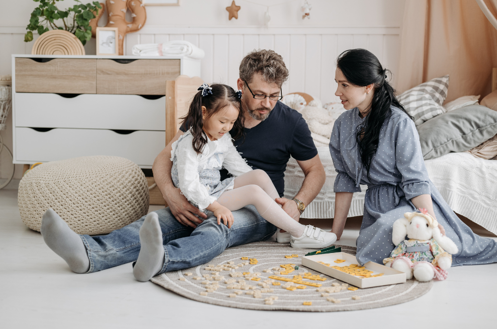 Parents and a child sitting on a carpet on the ground in the child's room, looking at a book.